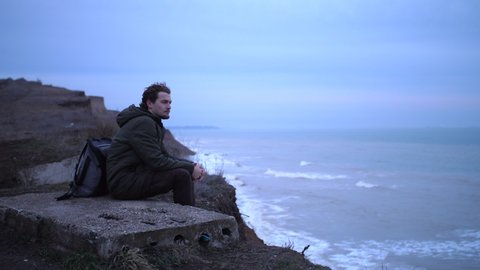 Sad lonely young man, with backpack, sits on a cement block at the edge of cliff above storm sea, and looks in a far lost in thought. Mist adds a moody atmosphere in late autumn cloudy day