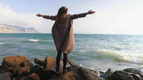 A woman stands on a rock by the sea and spread her arms out to the sides. Feels free. Enjoying the beautiful sea view. The concept of freedom.