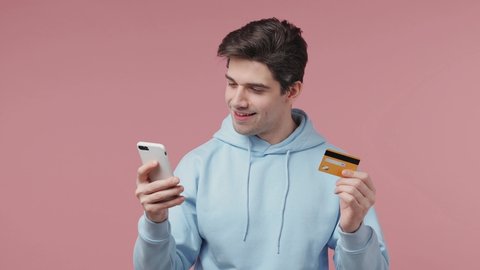Handsome young man 20s wears blue streetwear hoodie using mobile cell phone hold in hand credit bank card doing online shopping order booking tour isolated on pastel pink background studio portrait
