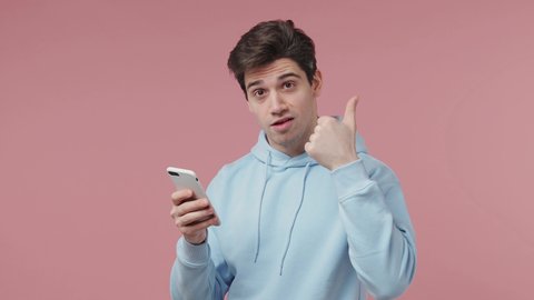 Young man 20s wears blue streetwear hoodie hold use mobile cell phone typing browsing chatting send sms doing online shopping order booking tour isolated on pastel pink wall background studio portrait