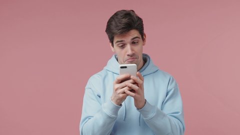 Young man 20s wears blue streetwear hoodie hold use mobile cell phone typing browsing chatting send sms doing online shopping order booking tour isolated on pastel pink wall background studio portrait