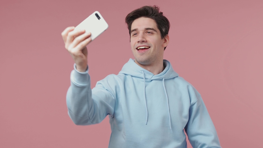 Handsome fun stylish young man 20s wears blue streetwear hoodie posing doing selfie shot on mobile phone post photo on social network internet isolated on pastel pink wall background studio portrait