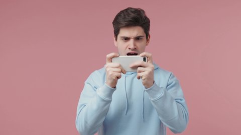 Gambling young man in blue streetwear hoodie use play racing on mobile cell phone hold gadget smartphone for pc video games isolated on pastel pink background studio People emotions lifestyle concept