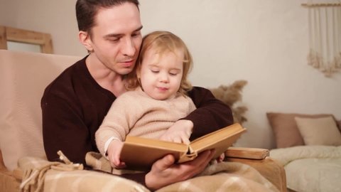 young father and daughter read a book, have fun, laugh. Home family evening, comfort and love. A man and a girl sit in a rocking chair, beige interior. close up