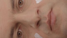 A young man rubs skin cream on his face on a blue background.Concept of applying skin cream to very close-up male face.Video for the vertical story.