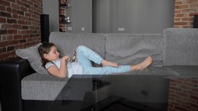 A little girl lies at home on the couch and looks into a smartphone. The child plays and watches the video. The girl has bare feet. The child is wearing pajamas. 4K