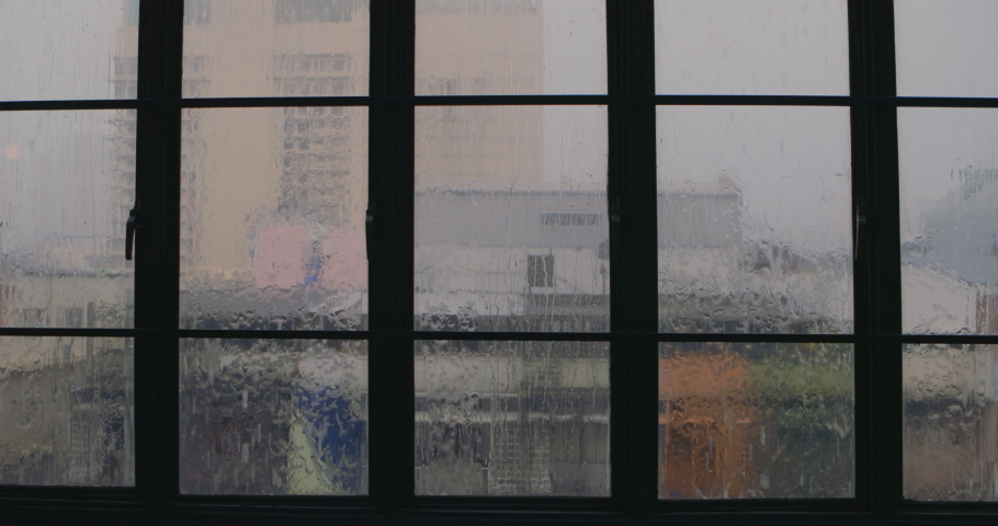 The interior of cute city apartment with large panoramic windows in the rain. Rain droplets on glass. Rain covered window with blurry city scape. Modern Asian city with skyscrapers in the background. Royalty-Free Stock Footage #1070106787