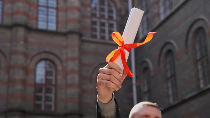 Male hand raised up holding diploma of academy graduate on the background of beautiful university school building Education concept Young graduated guy holding graduation degree convocation ceremony Royalty-Free Stock Footage #1070121046