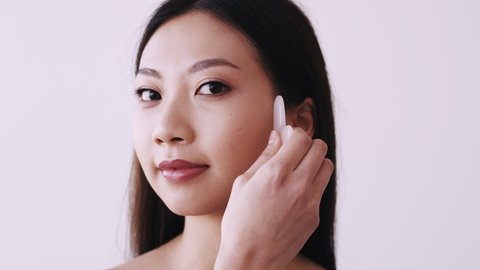 Gua sha massage. Asian skin care. Acupressure therapy. Alternative beauty procedure. Portrait of Chinese woman with nude makeup touching face with pink rose quartz isolated on light background.