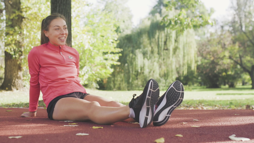 Fitness girl stretching her legs while sitting on an all weather synthetic track surface in the park. Warming up exercise for running.