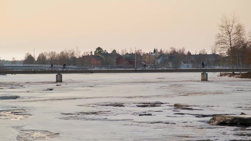 People Walking and Cycling on a Bridge over a Frozen River with Beautiful Old Wooden Houses in the Background in Winter