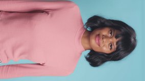 Vertical video happy young dark skinned lovely woman broadly smiles satisfied expression look at camera poses amused listen funny story dressed in pink jumper indoor over blue background. Joy concept