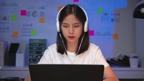 Cheerful beautiful Asian woman in a white t-shirt with wearing headphones and look to laptop at video calling meeting and study online on the Internet.
