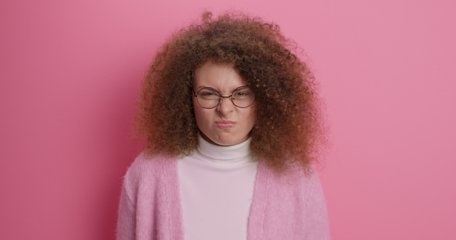 Outraged curly haired young woman looks angrily at camera sakes hands and head expresses negative emotions wears casual jumper stands irritated against pink background has argument with someone
