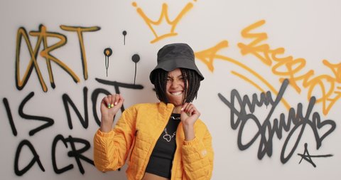 Young fashionable graffiti artist dances carefree wears black hat and yellow jacket dances to favorite music poses near graffiti wall has fun. Youth culture lifestyle and contemporary art concept