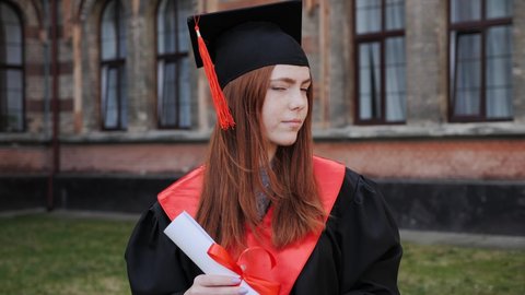 Portrait of a beautiful ginger female student dressed in a mantle and caps with a master's degree in hand university graduate on the background of a high school academy graduation day.