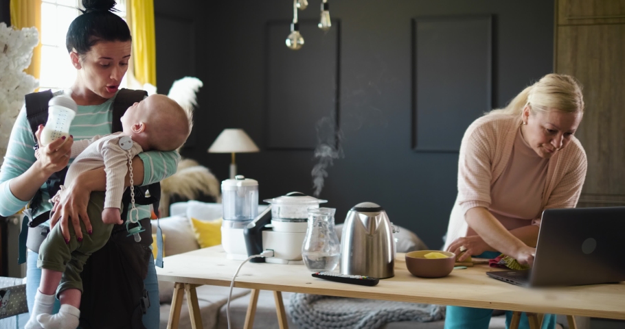 Young mother feeds the baby in a baby carrier, the grandmother watches tutorials for cooking food on a steam cooker