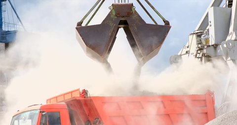 Grapple crane unloading cement clinker from ship to truck in port. Grab is discharging bulk cargo from a hold of cargo ship, causing dust in atmosphere. Loading in Harbor outdoors. Logistics 