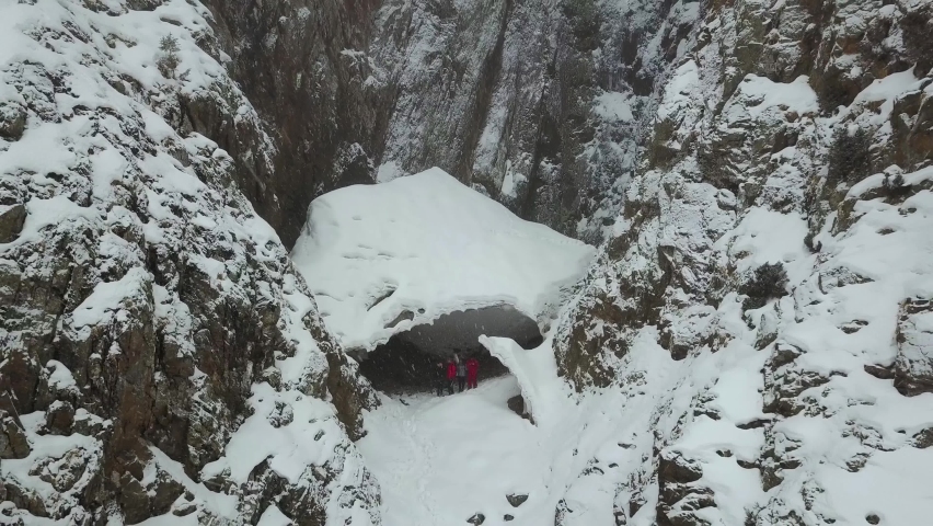 Ice cave in the mountains. View from a drone. It is snowing, and the gorge is covered with fog. A group of people in a cave. The view from the top. Spruce trees grow on large rocks. Near the forest.