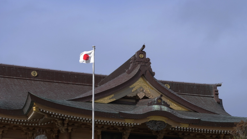 Japanese flag and the roof of shrine. Flag is waving with wind. Traditionally decorated architecture. Blue sky background. Kabushima Shrine in Hachinohe, Aomori, Tohoku, Japan. Crane family crest