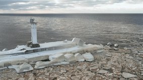 Beautiful aerial view of a snowy frozen lighthouse and pier surrounded by broken ice floes floating in sea. Cloudy blue sky in the background. Backward motion. 4k footage.