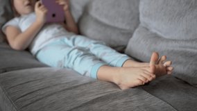 A young girl lies at home on the couch and looks into a pink tablet. Focus on the feet. The child is dressed in pajamas and has bare feet. Close up. 4K