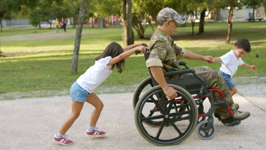 Kids helping disabled military dad to wheel heavy wheelchair. Ex soldier walking with children in park. Veteran of war or returning home concept
