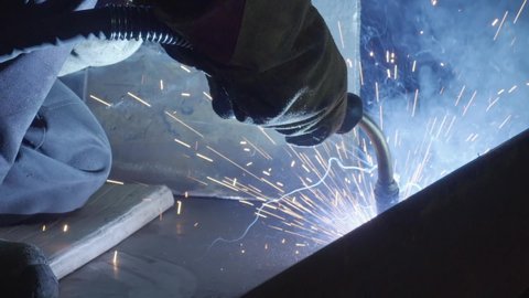 Slow-Motion. Turbine Production Plant. A Worker in Protective Clothing and a Helmet is Holding a Welding Torch against a Part of the Turbine. Sparks are Flying Out. Welding Process.