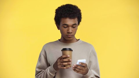 african american teenager drinking and using smartphone isolated on yellow