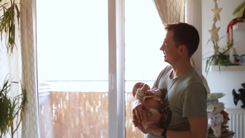 Young dad makes movements with child in arms near window apartment. It rocks baby to sleep. Small child care. Contact between newborn and father. Confident behavior with the child.