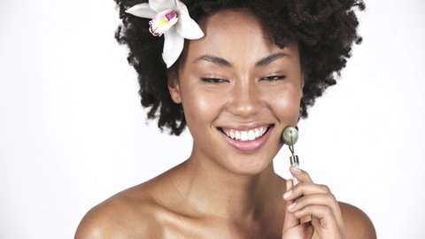 attractive naked african american woman massaging clean face with jade roller and smiling on white