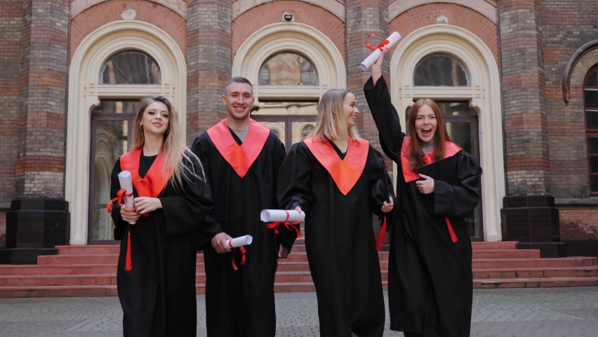 A group of academy graduates in gowns and certificates with honors go to celebrate graduation ceremony on the background of the university. Young smiling people laughing having fun Higher education.