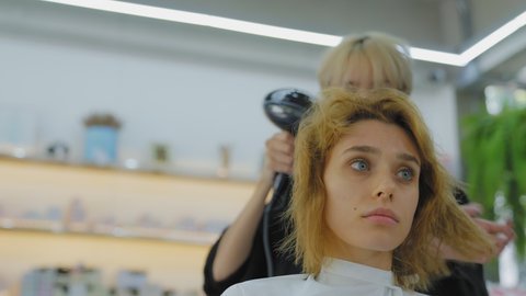 Professional woman Asian barber, after cutting a caucasian, white European clientele, blow her hair with a hair dryer in a modern beauty salon.