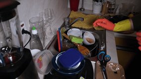 Closeup fast motion video of a young housewife in orange rubber gloves washing dishes in the very messy kitchen. Colorful dirty plates, bowls and mugs everywhere. Chaotic, real kitchen scene. 4k