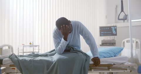 Portrait of afro-american sick patient sitting on hospital bed and rubbing face feeling unwell. Tired and exhausted sick black man sitting in hospital ward