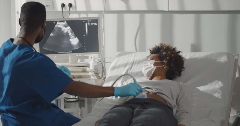 Young afro-american pregnant woman in safety at ultrasound office in modern clinic. Portrait of young male doctor wearing safety mask and gloves using ultrasound machine to check pregnant woman. Royalty-Free Stock Footage #1070187433