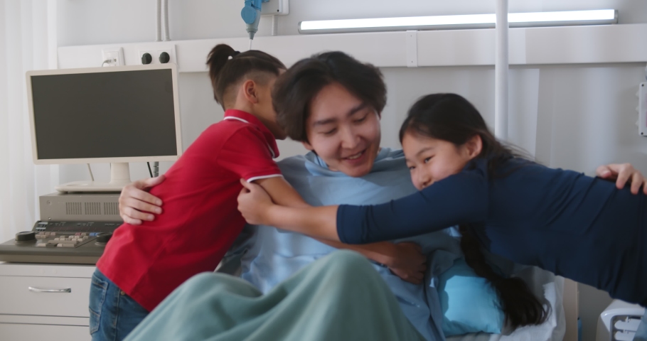 Diverse children visiting adoptive asian father in hospital ward. Sick korean man resting in hospital bed and hugging kids visiting in clinic room. Healthcare and family concept