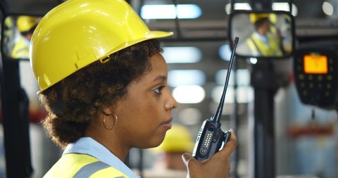 Side view portrait of afro-american woman sitting in forklift and using walkie-talkie while moving goods in warehouse. Close up of forklift female driver communicating over portable radio