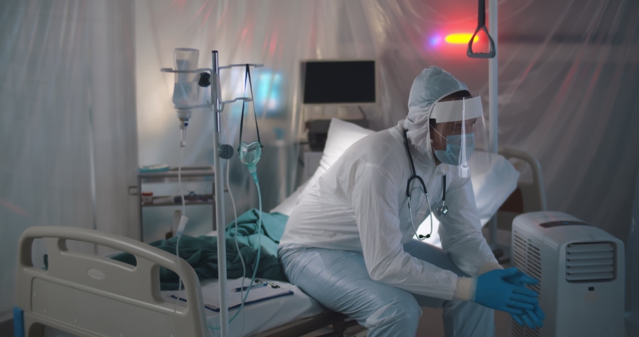 Sad tired doctor in protective suit and medical mask sitting on empty bed in hospital ward. Exhausted and stressed medical worker in ppe suit resting on bed in quarantine room Royalty-Free Stock Footage #1070187484