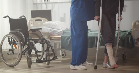 Cropped shot of doctor helping patient with bandaged leg walking on crutches in hospital ward. Traumatologist assisting patient with broken leg in clinic