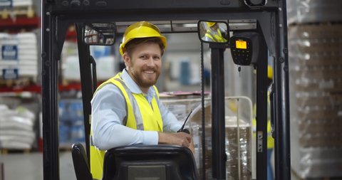 Portrait young male warehouse worker sitting in forklift truck and smiling at camera. Happy forklift driver looking at camera working in large industrial storehouse