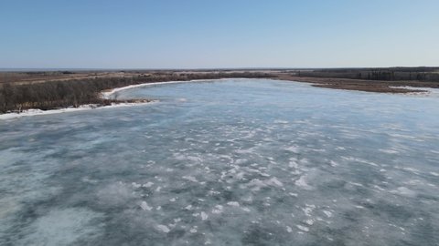 Drone flying slowly forward over a frozen lake with the blue sky reflecting on the ice. A bald eagle can be seen in the distance sitting on the ice.
