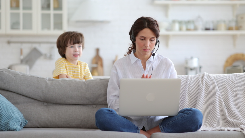 Mother freelancer talking on video call, remote working on laptop from home office with kid during lockdown. Children make noise disturb woman at work, shows tongue standing behind couch. Distance job Royalty-Free Stock Footage #1070197588