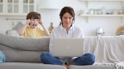 Mother freelancer talking on video call, remote working on laptop from home office with kid during lockdown. Children make noise disturb woman at work, shows tongue standing behind couch. Distance job