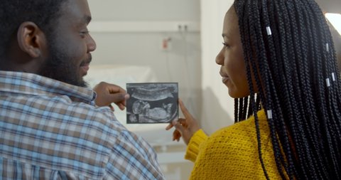 Back view of young married african couple looking at ultrasound picture at clinic. Happy black husband and wife sitting in hospital and holding sonogram picture