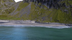 Drone footage flying inland over the sea towards a beautiful sandy beach and green mountainside. Kvalvika Beach, a famous tourist hiking hotspot in Lofoten, Norway.