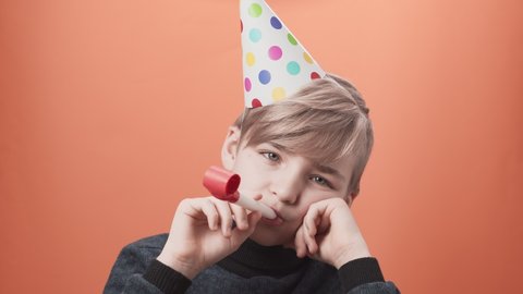 Young sad schoolboy in birthday hat blowing a whistle on orange background. Lonely upset birthday kid at quarantine time.