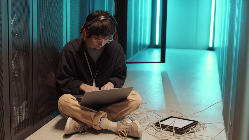 Wide shot of young Asian IT technician wearing casual clothes and headphones sitting on floor of server room and coding on laptop