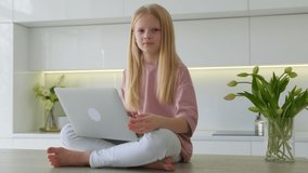Caucasian blonde child schoolgirl girl looks at camera while sitting at laptop and is engaged in online learning from home watching web class lesson or listening to tutor on video call.