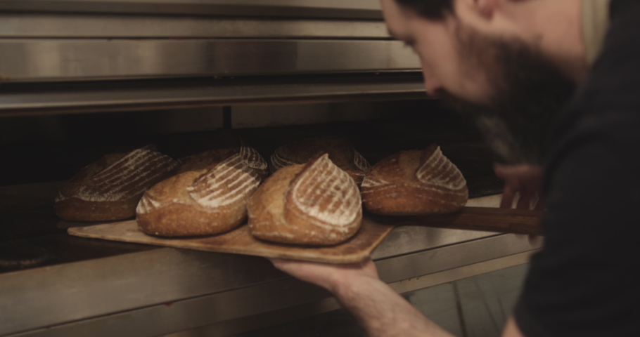 Bearded male baker taking baked sourdough bread out of the oven wearing apron in bakery | Shutterstock HD Video #1070215468